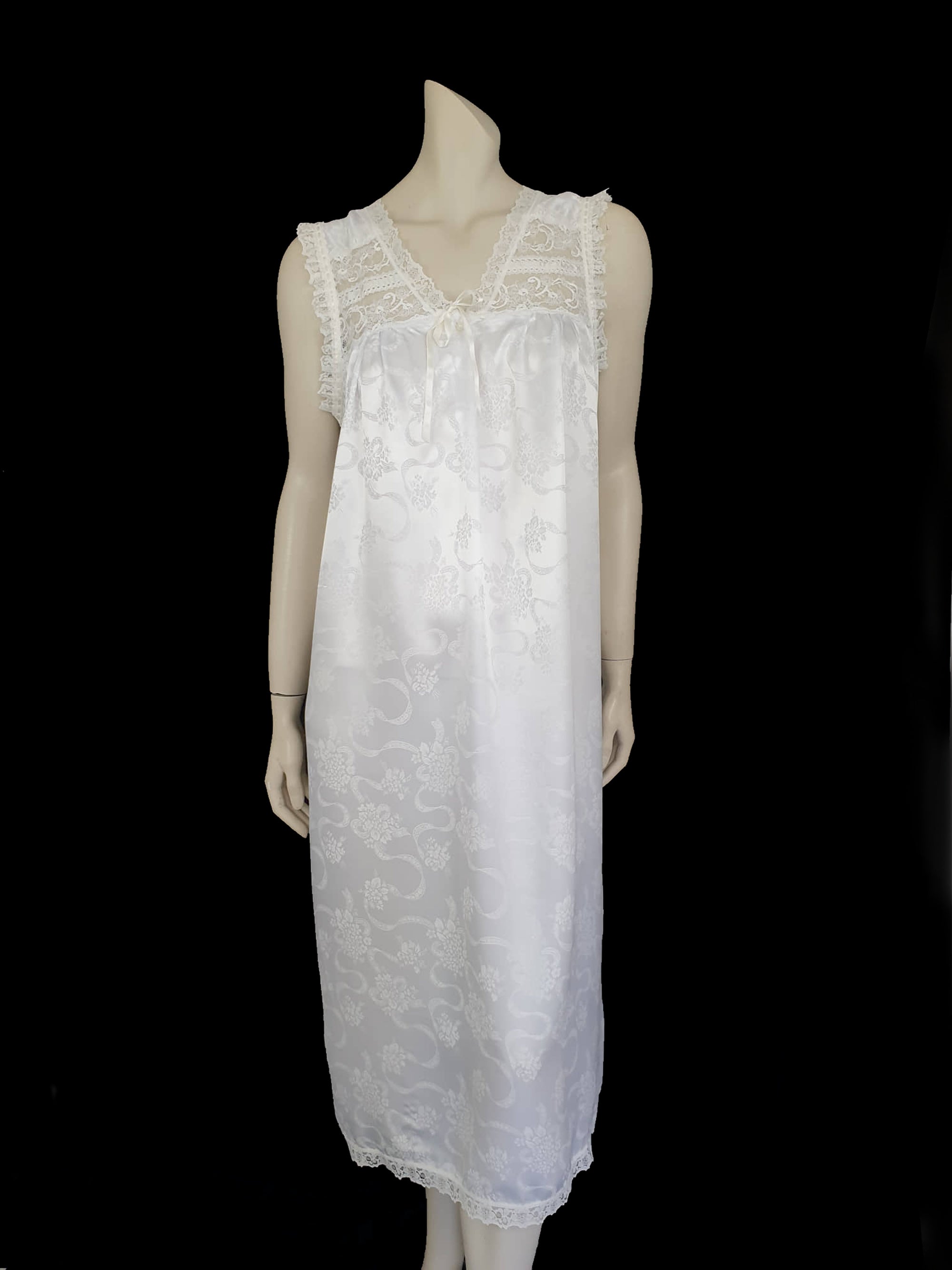 White satin nightgown has a lacy yoke, V-neck and has lace trim at the neckline, armholes and hem. The beautiful fabric is a damask weave with a floral design. Closes at the neck with one button and a ribbon bow. The nightie is mid to lower calf length. Fabric is polyester. Made in Australia by Texwear - 1980s. Medium