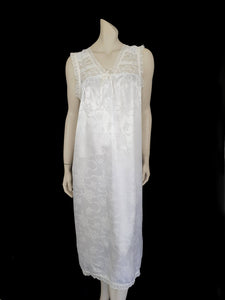 White satin nightgown has a lacy yoke, V-neck and has lace trim at the neckline, armholes and hem. The beautiful fabric is a damask weave with a floral design. Closes at the neck with one button and a ribbon bow. The nightie is mid to lower calf length. Fabric is polyester. Made in Australia by Texwear - 1980s. Medium