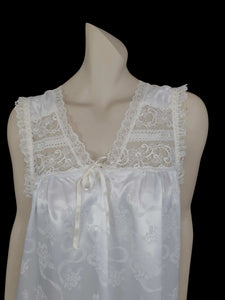 White satin nightgown has a lacy yoke, V-neck and has lace trim at the neckline, armholes and hem. The beautiful fabric is a damask weave with a floral design. Closes at the neck with one button and a ribbon bow. The nightie is mid to lower calf length. Fabric is polyester. Made in Australia by Texwear - 1980s. Medium