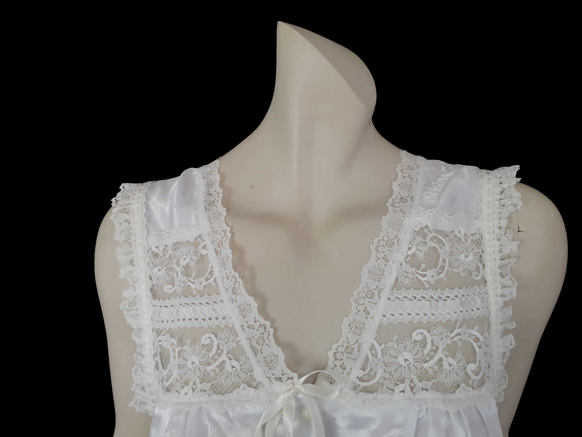 White satin nightgown has a lacy yoke, V-neck and has lace trim at the neckline, armholes and hem. The beautiful fabric is a damask weave with a floral design. Closes at the neck with one button and a ribbon bow. The nightie is mid to lower calf length. Fabric is polyester. Made in Australia by Texwear - 1980s. Medium