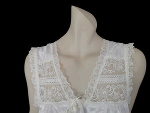 White satin nightgown has a lacy yoke, V-neck and has lace trim at the neckline, armholes and hem. The beautiful fabric is a damask weave with a floral design. Closes at the neck with one button and a ribbon bow. The nightie is mid to lower calf length. Fabric is polyester. Made in Australia by Texwear - 1980s. Medium