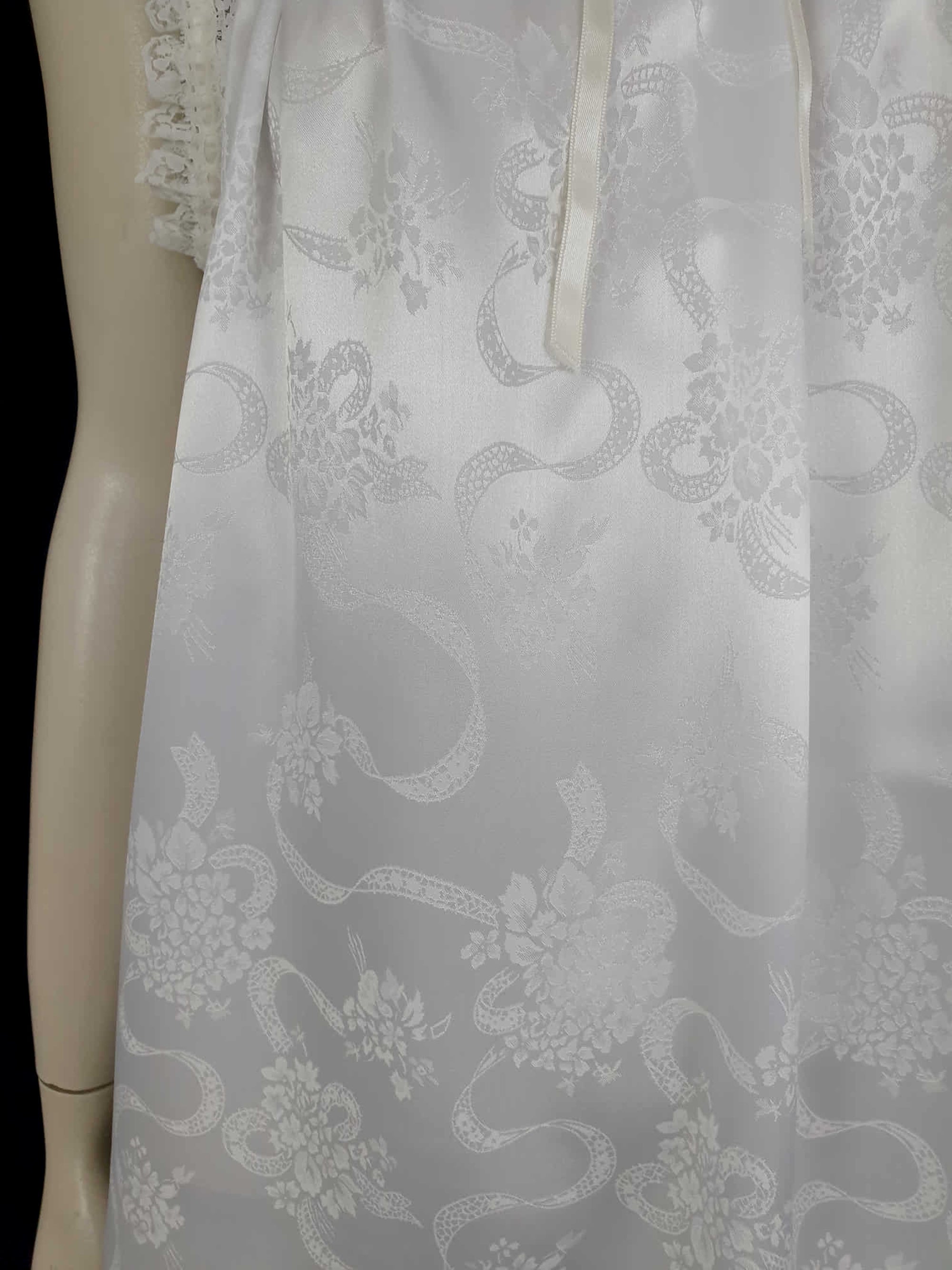 White satin nightgown has a lacy yoke, V-neck and has lace trim at the neckline, armholes and hem. The beautiful fabric is a damask weave with a floral design. Closes at the neck with one button and a ribbon bow. The nightie is mid to lower calf length. Fabric is polyester. Made in Australia by Texwear - 1980s. Medium