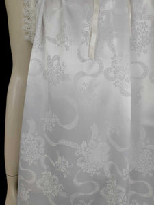 White satin nightgown has a lacy yoke, V-neck and has lace trim at the neckline, armholes and hem. The beautiful fabric is a damask weave with a floral design. Closes at the neck with one button and a ribbon bow. The nightie is mid to lower calf length. Fabric is polyester. Made in Australia by Texwear - 1980s. Medium