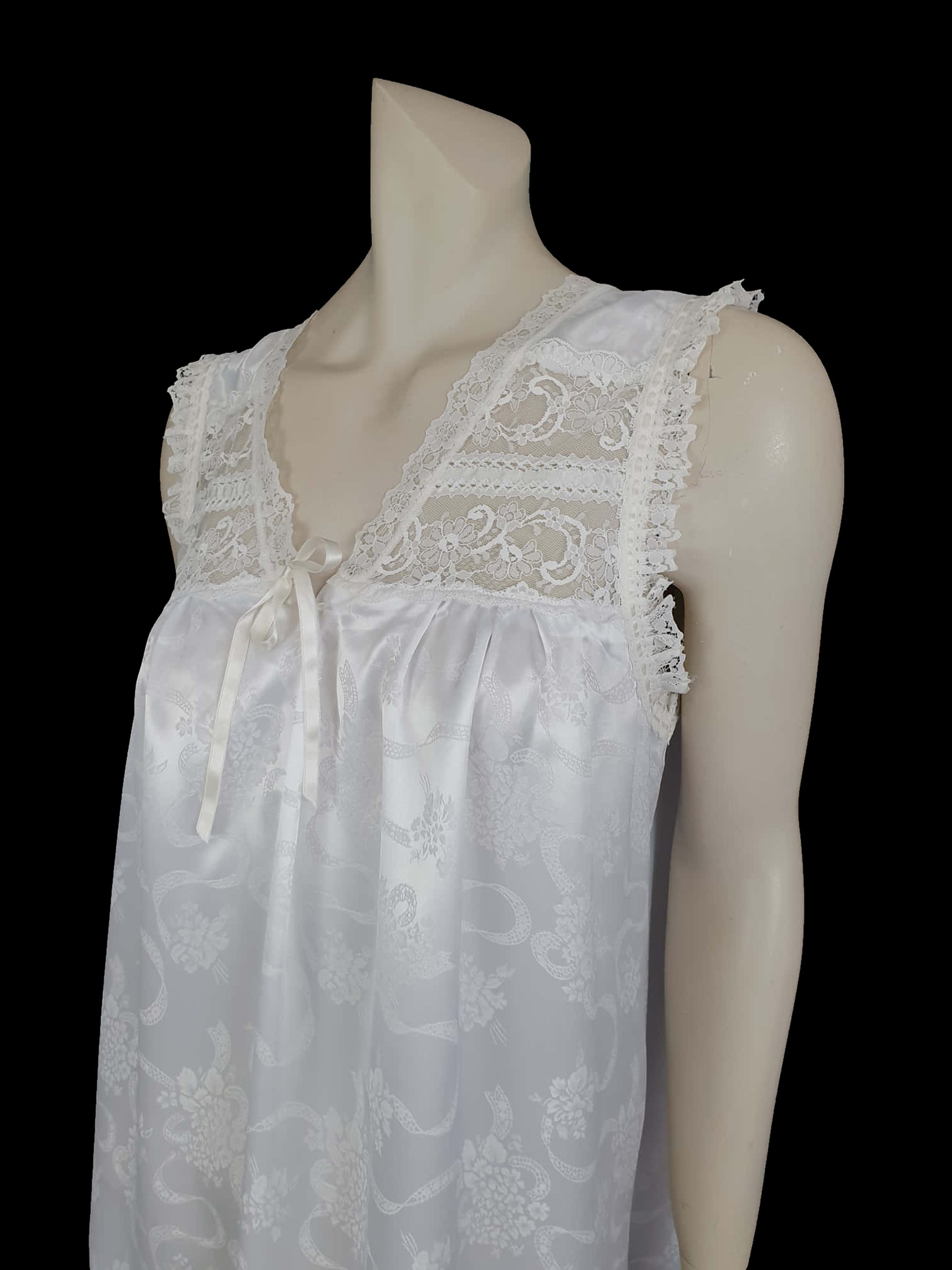 White satin nightgown has a lacy yoke, V-neck and has lace trim at the neckline, armholes and hem. The beautiful fabric is a damask weave with a floral design. Closes at the neck with one button and a ribbon bow. The nightie is mid to lower calf length. Fabric is polyester. Made in Australia by Texwear - 1980s. Medium
