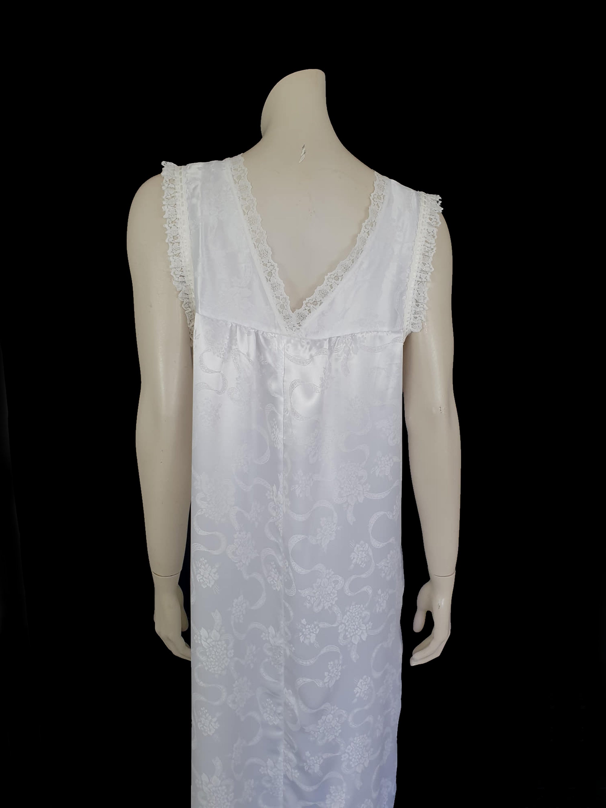 White satin nightgown has a lacy yoke, V-neck and has lace trim at the neckline, armholes and hem. The beautiful fabric is a damask weave with a floral design. Closes at the neck with one button and a ribbon bow. The nightie is mid to lower calf length. Fabric is polyester. Made in Australia by Texwear - 1980s. Medium