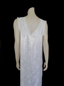 White satin nightgown has a lacy yoke, V-neck and has lace trim at the neckline, armholes and hem. The beautiful fabric is a damask weave with a floral design. Closes at the neck with one button and a ribbon bow. The nightie is mid to lower calf length. Fabric is polyester. Made in Australia by Texwear - 1980s. Medium