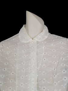 Beautiful sheer white nylon blouse embroidered all over in the eyelet style. 
The blouse has a peter pan collar and capped sleeves. It's darted in at the waist as it was designed to be worn with high waisted skirts. Closes to above waist level with clear plastic buttons. Fabric is nylon. No brand label. Late 1950s or very early 1960s. Medium