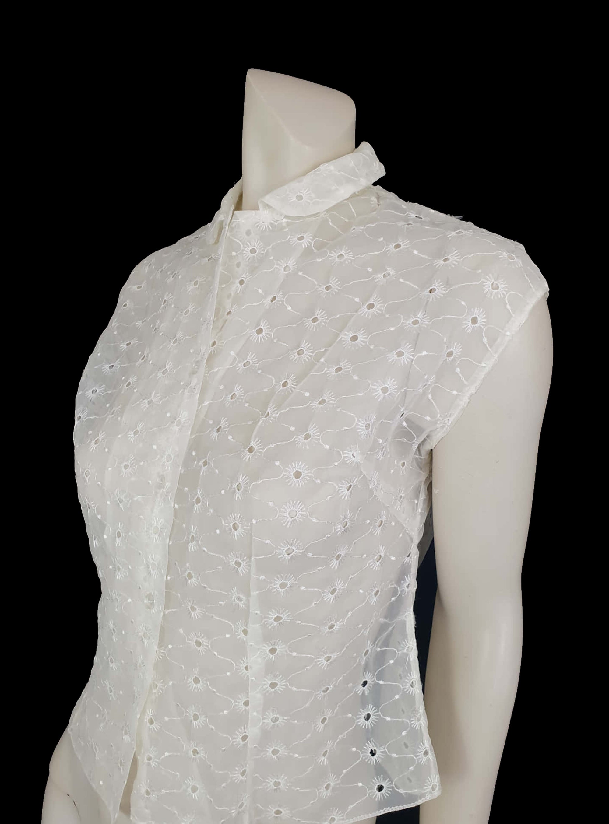 Beautiful sheer white nylon blouse embroidered all over in the eyelet style. 
The blouse has a peter pan collar and capped sleeves. It's darted in at the waist as it was designed to be worn with high waisted skirts. Closes to above waist level with clear plastic buttons. Fabric is nylon. No brand label. Late 1950s or very early 1960s. Medium