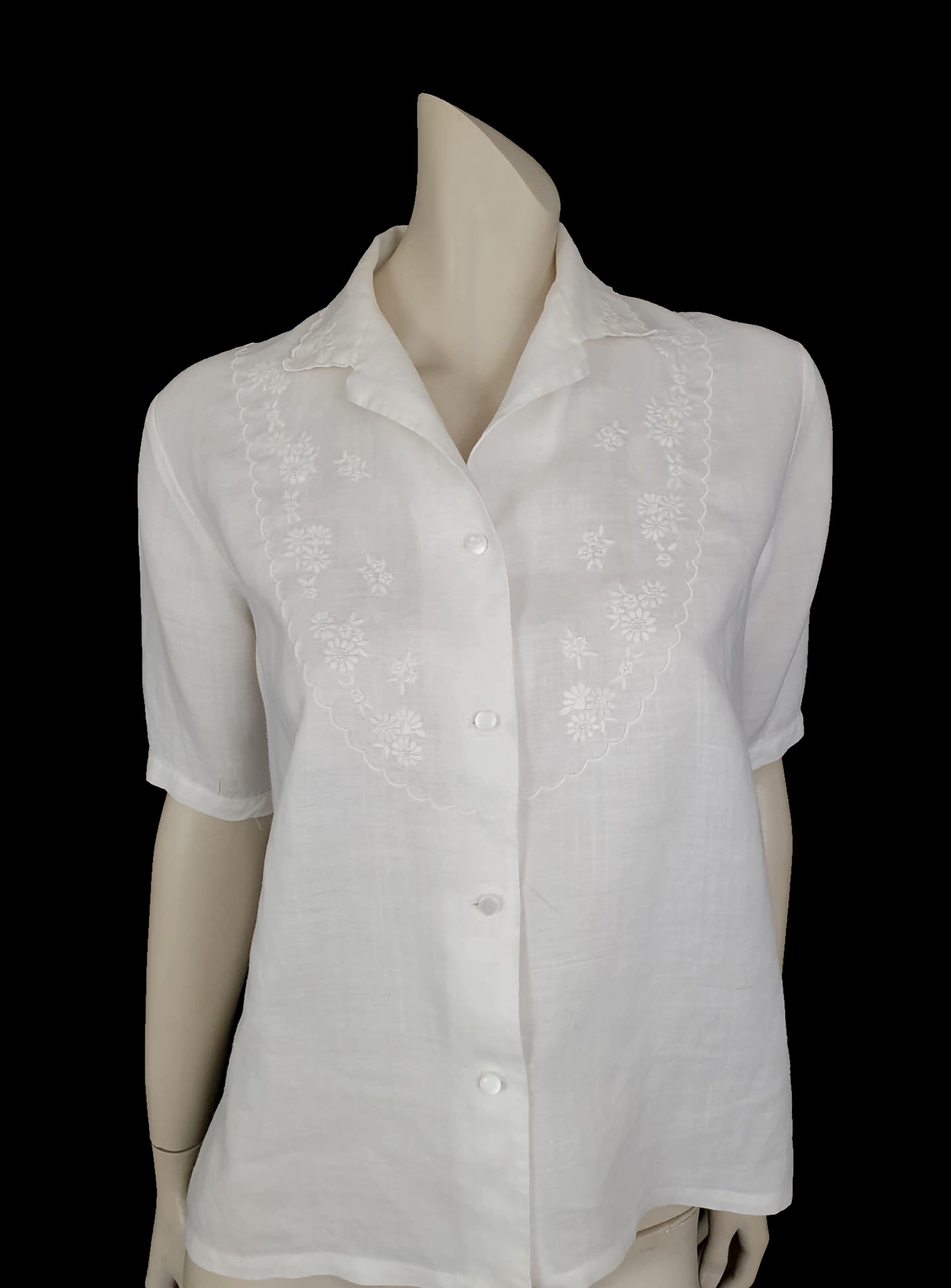 White linen blouse has flowers embroidered over the front and collar. The small collar has a scalloped embroidered edging. Above-elbow length sleeves are finished with a band and close with a button. Button front closure. Made in China by Robert Lim International - 1990s. Medium