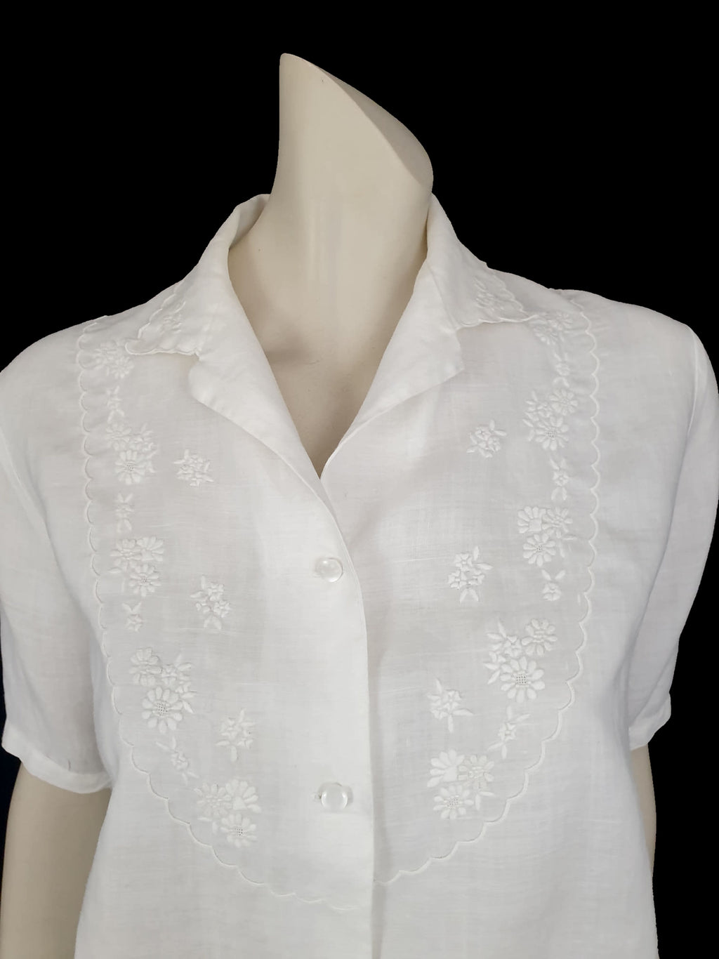 White linen blouse has flowers embroidered over the front and collar. The small collar has a scalloped embroidered edging. Above-elbow length sleeves are finished with a band and close with a button. Button front closure. Made in China by Robert Lim International - 1990s. Medium