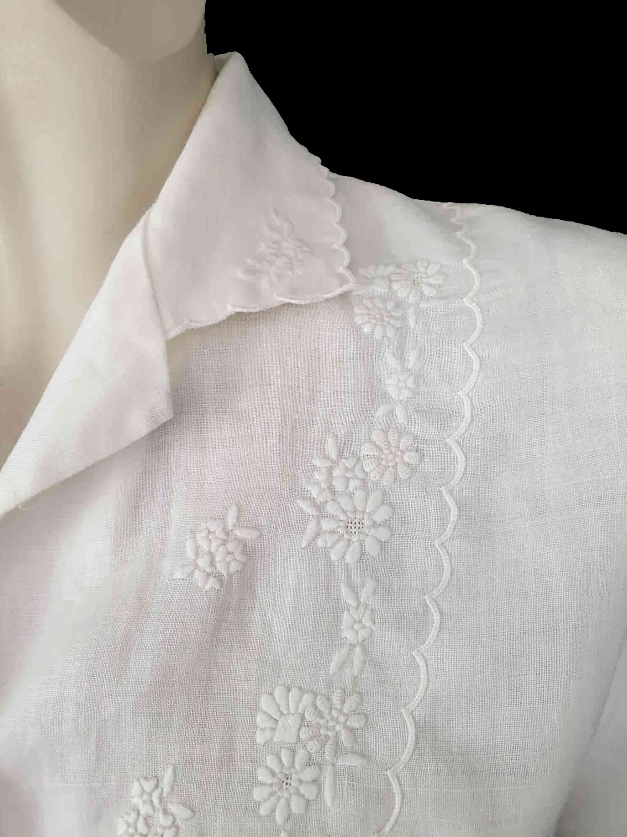 White linen blouse has flowers embroidered over the front and collar. The small collar has a scalloped embroidered edging. Above-elbow length sleeves are finished with a band and close with a button. Button front closure. Made in China by Robert Lim International - 1990s. Medium