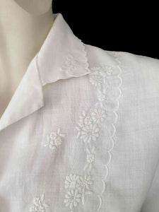 White linen blouse has flowers embroidered over the front and collar. The small collar has a scalloped embroidered edging. Above-elbow length sleeves are finished with a band and close with a button. Button front closure. Made in China by Robert Lim International - 1990s. Medium