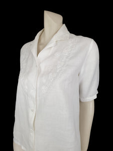 White linen blouse has flowers embroidered over the front and collar. The small collar has a scalloped embroidered edging. Above-elbow length sleeves are finished with a band and close with a button. Button front closure. Made in China by Robert Lim International - 1990s. Medium