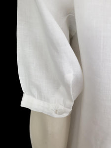 White linen blouse has flowers embroidered over the front and collar. The small collar has a scalloped embroidered edging. Above-elbow length sleeves are finished with a band and close with a button. Button front closure. Made in China by Robert Lim International - 1990s. Medium