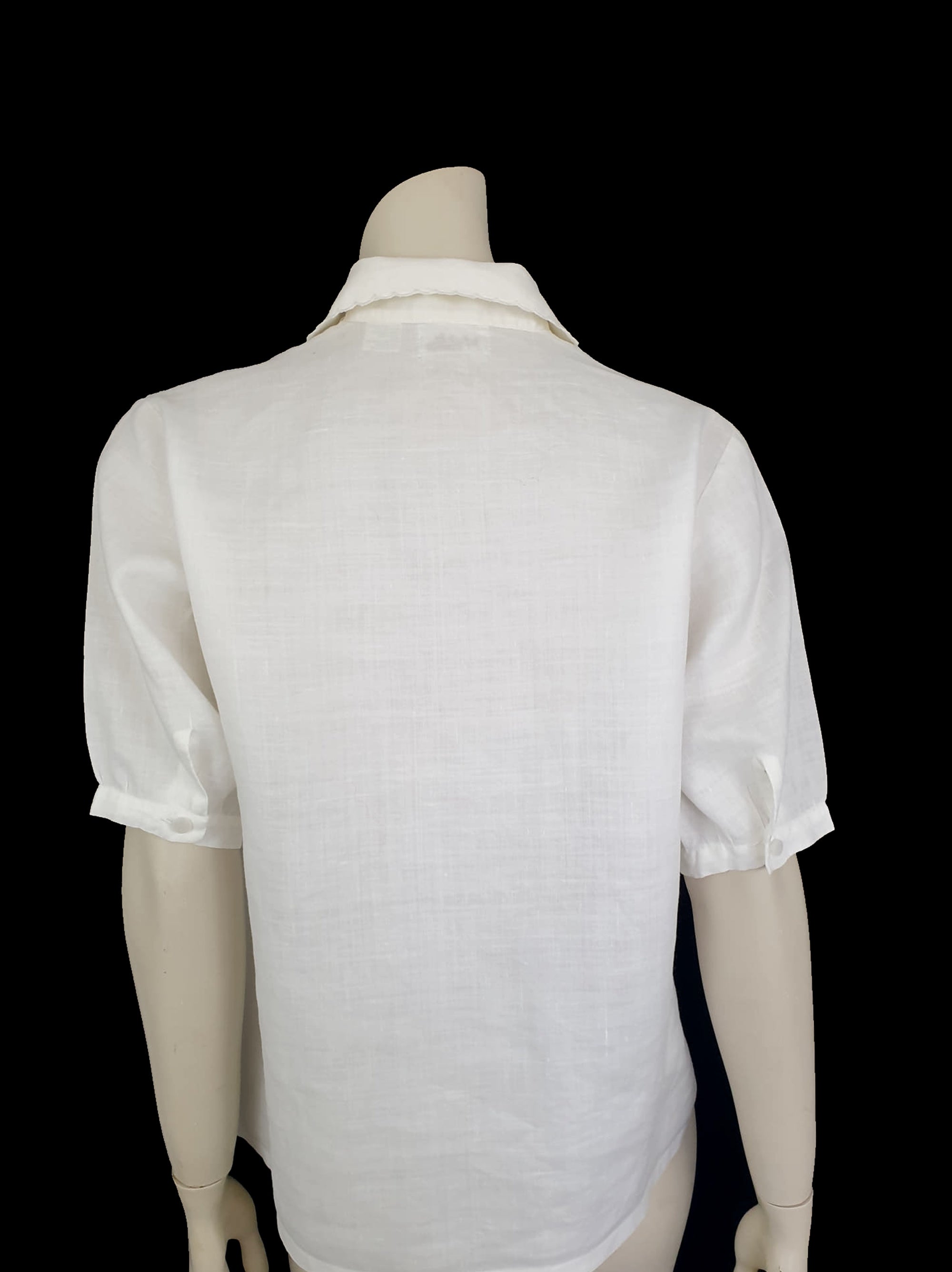 White linen blouse has flowers embroidered over the front and collar. The small collar has a scalloped embroidered edging. Above-elbow length sleeves are finished with a band and close with a button. Button front closure. Made in China by Robert Lim International - 1990s. Medium