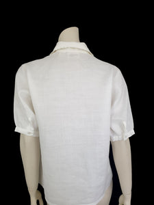 White linen blouse has flowers embroidered over the front and collar. The small collar has a scalloped embroidered edging. Above-elbow length sleeves are finished with a band and close with a button. Button front closure. Made in China by Robert Lim International - 1990s. Medium