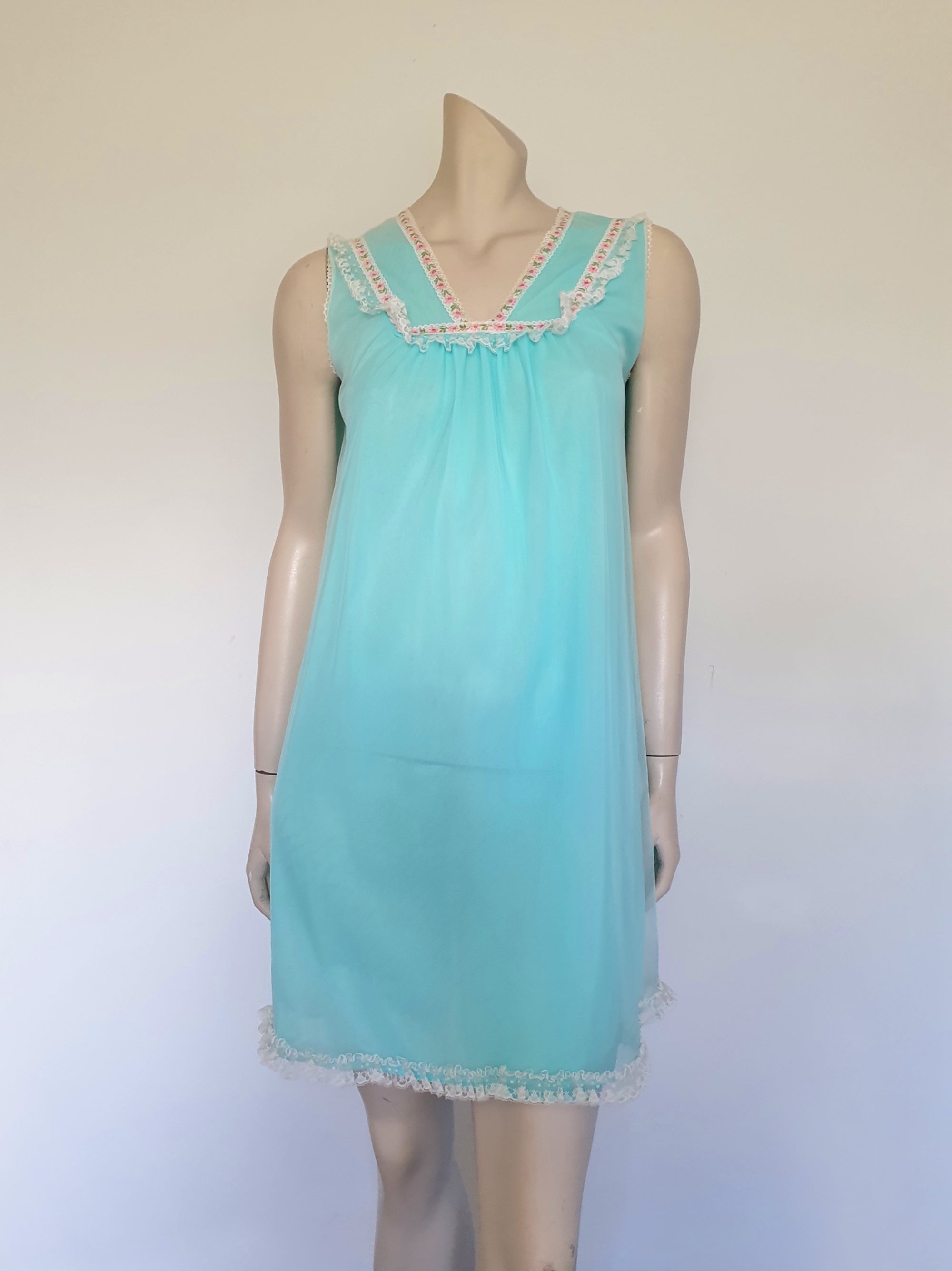 Very cute little aqua mini nightgown with floral braid and lace trim at the V neckline. The same lace also trims the hem. The nightie has a sheer overlay with an opaque layer of fabric beneath. Fabric is nylon. Brand is Irene Lingerie - Late 1960s - 1970s. Small.
