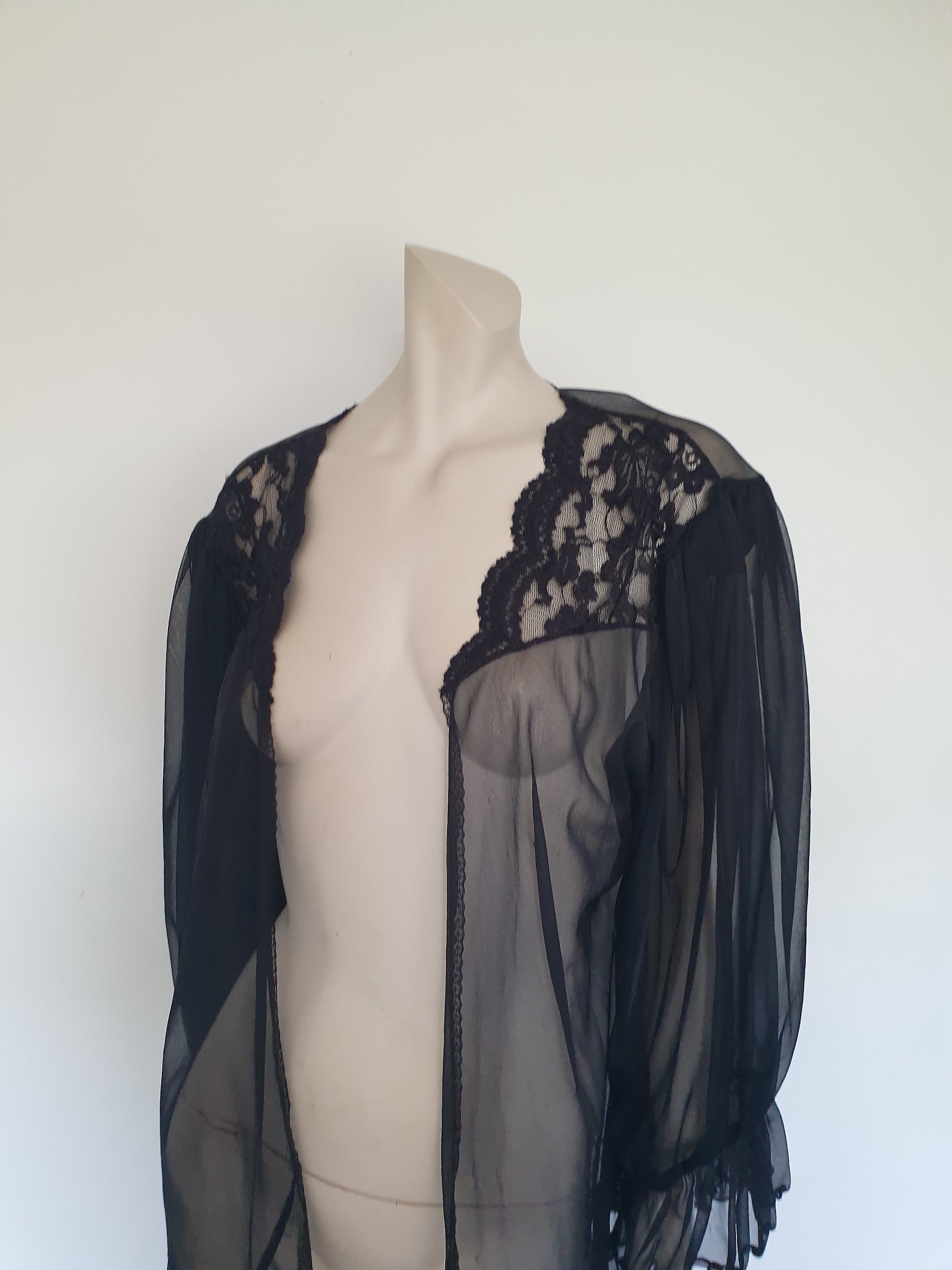 Sheer black mini length robe has long, gathered sleeves with sleeve headers and elastic at the wrists. Lace panels at the front yoke and ruffled trim. Narrow lace trim at the front opening and neckline. No closure. Fabric is nylon. Label says S. G. for Escante. 1980s-1990s. Extra Large.