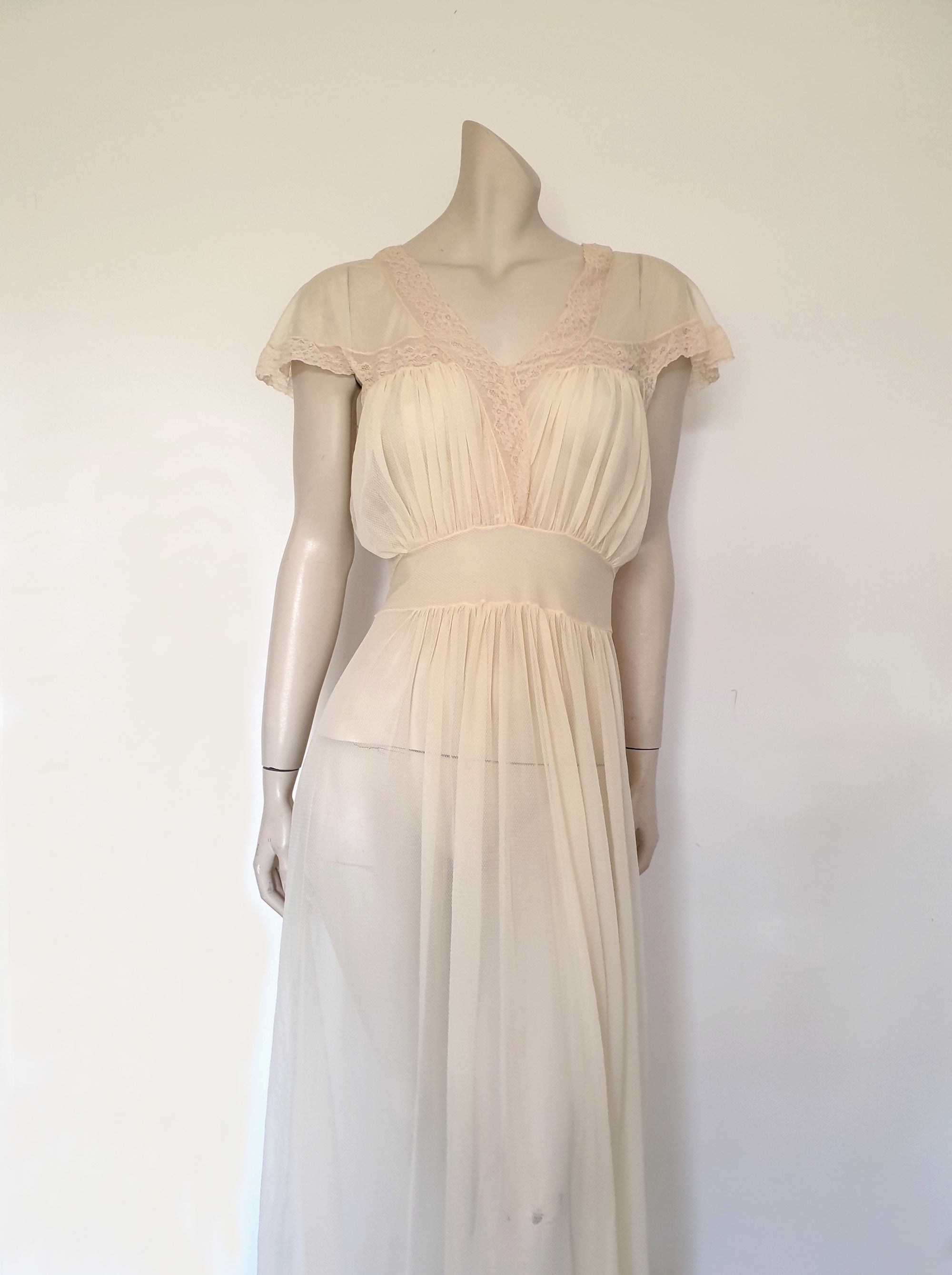 Glorious sheer cream nightgown for sexy '50s glamour. Beautiful gown is gathered over the bust into panels of wide ecru cotton lace which extend across into capped sleeves and a V neckline. The neckline at the back is squared off. Bodice and skirt are gathered into the midriff yoke. The skirt is flared and a tie extends from the side seams. The nylon fabric has a pattern in the weave and is quite sheer. Brand is Prestige - 1950s. Small