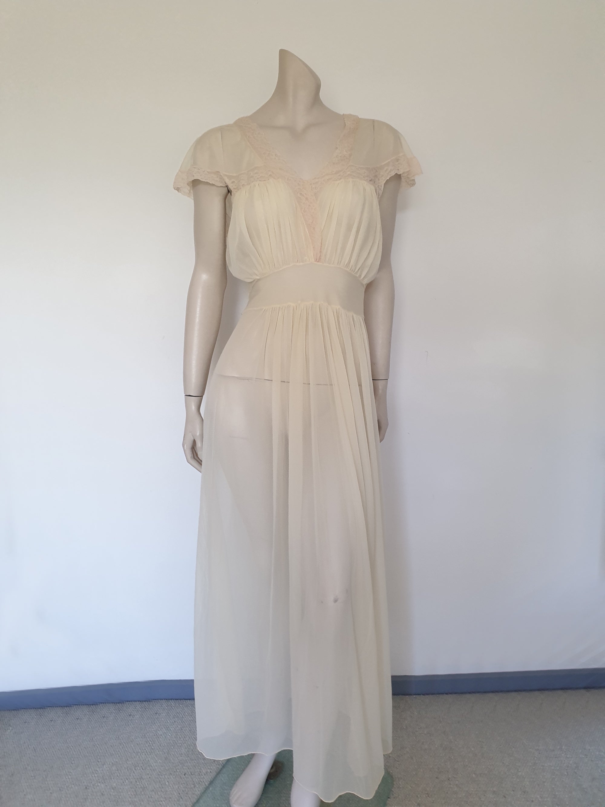 Glorious sheer cream nightgown for sexy '50s glamour. Beautiful gown is gathered over the bust into panels of wide ecru cotton lace which extend across into capped sleeves and a V neckline. The neckline at the back is squared off. Bodice and skirt are gathered into the midriff yoke. The skirt is flared and a tie extends from the side seams. The nylon fabric has a pattern in the weave and is quite sheer. Brand is Prestige - 1950s. Small
