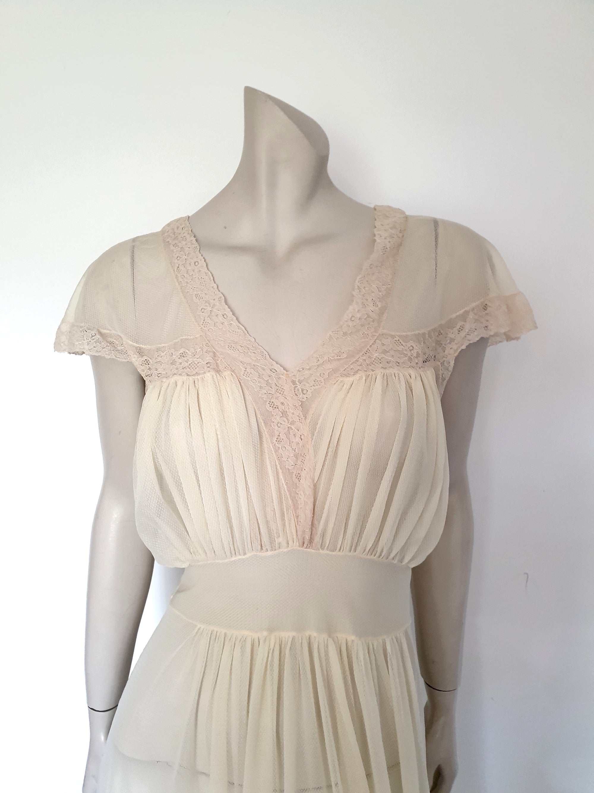 Glorious sheer cream nightgown for sexy '50s glamour. Beautiful gown is gathered over the bust into panels of wide ecru cotton lace which extend across into capped sleeves and a V neckline. The neckline at the back is squared off. Bodice and skirt are gathered into the midriff yoke. The skirt is flared and a tie extends from the side seams. The nylon fabric has a pattern in the weave and is quite sheer. Brand is Prestige - 1950s. Small