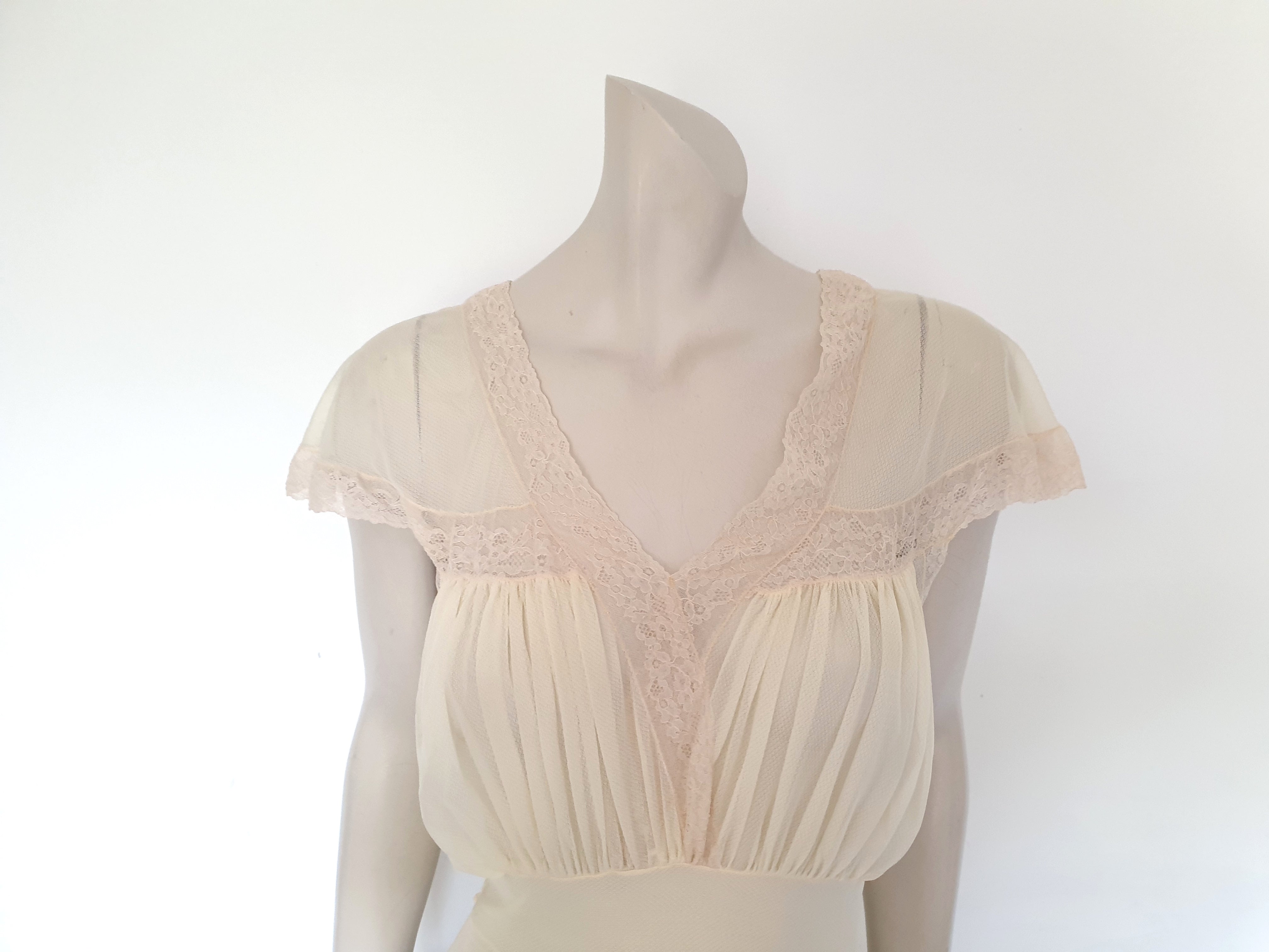 Glorious sheer cream nightgown for sexy '50s glamour. Beautiful gown is gathered over the bust into panels of wide ecru cotton lace which extend across into capped sleeves and a V neckline. The neckline at the back is squared off. Bodice and skirt are gathered into the midriff yoke. The skirt is flared and a tie extends from the side seams. The nylon fabric has a pattern in the weave and is quite sheer. Brand is Prestige - 1950s. Small