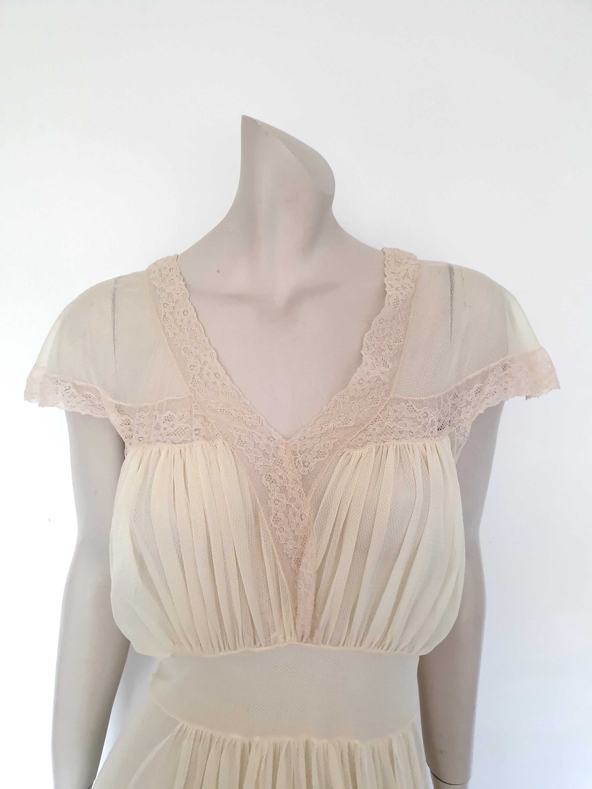 Glorious sheer cream nightgown for sexy '50s glamour. Beautiful gown is gathered over the bust into panels of wide ecru cotton lace which extend across into capped sleeves and a V neckline. The neckline at the back is squared off. Bodice and skirt are gathered into the midriff yoke. The skirt is flared and a tie extends from the side seams. The nylon fabric has a pattern in the weave and is quite sheer. Brand is Prestige - 1950s. Small