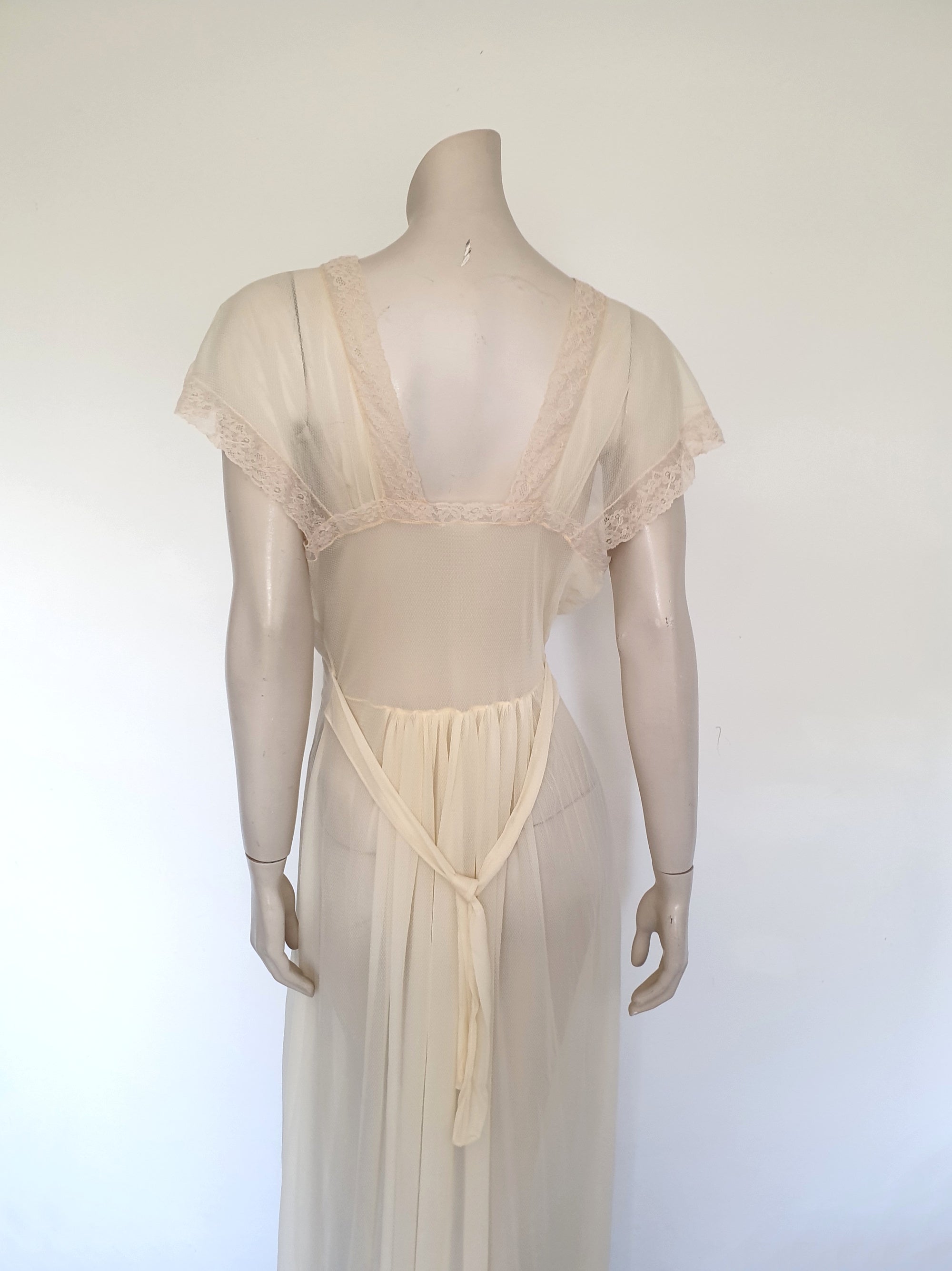 Glorious sheer cream nightgown for sexy '50s glamour. Beautiful gown is gathered over the bust into panels of wide ecru cotton lace which extend across into capped sleeves and a V neckline. The neckline at the back is squared off. Bodice and skirt are gathered into the midriff yoke. The skirt is flared and a tie extends from the side seams. The nylon fabric has a pattern in the weave and is quite sheer. Brand is Prestige - 1950s. Small