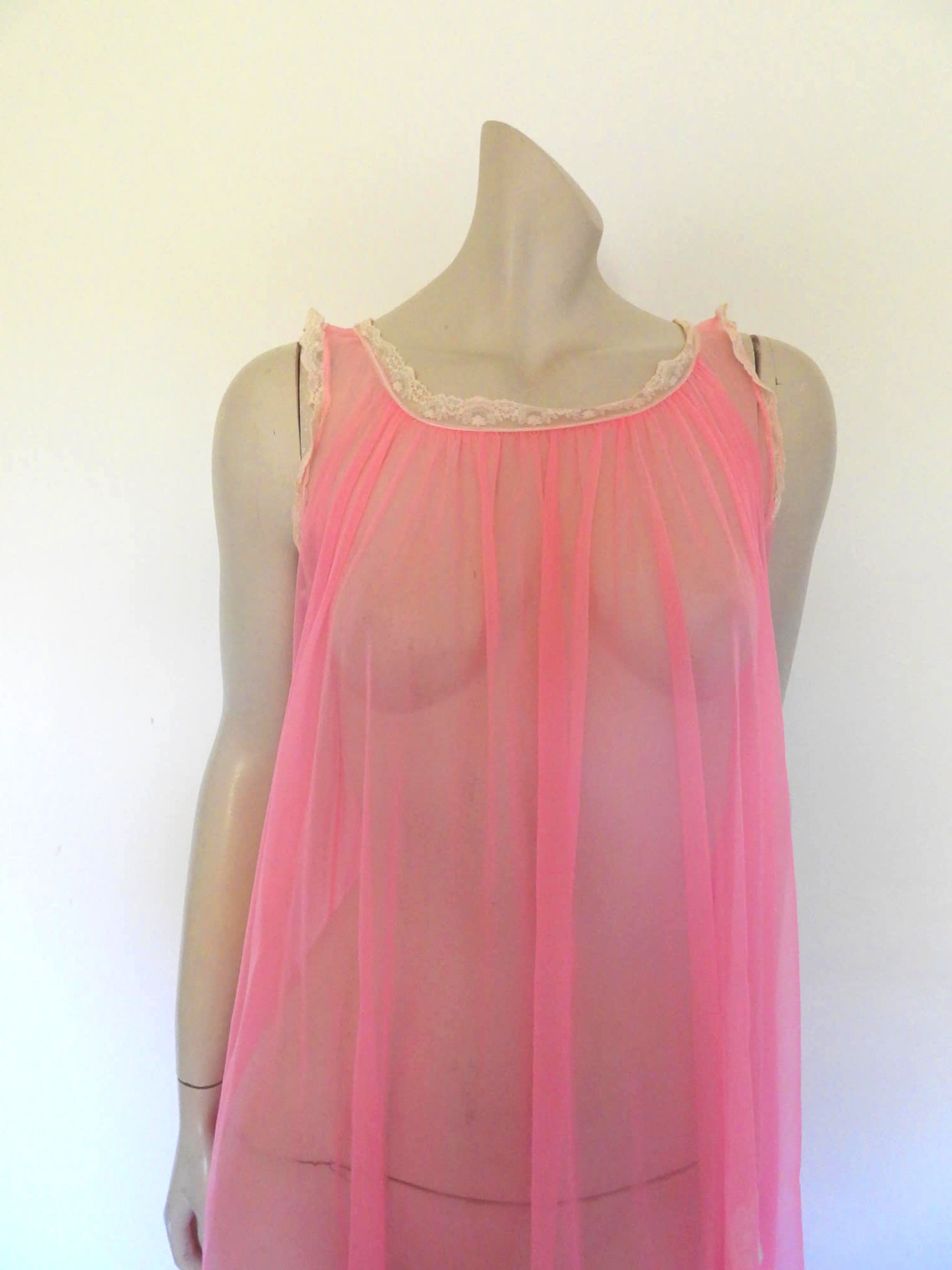 Super sexy sheer hot pink negligee nightgown. Has some issues but presents well. Very full negligee has wide neckline trimmed with cream lace. The hem is trimmed with a deep border of cream lace. There's a fixed tie at centre back which can tie in the fullness if you prefer. I have appliqued two flowers over two large holes in the front. Fabric is a single layer of sheer nylon. Made in the USA for Berkliff - 1960s. Small to Medium.
