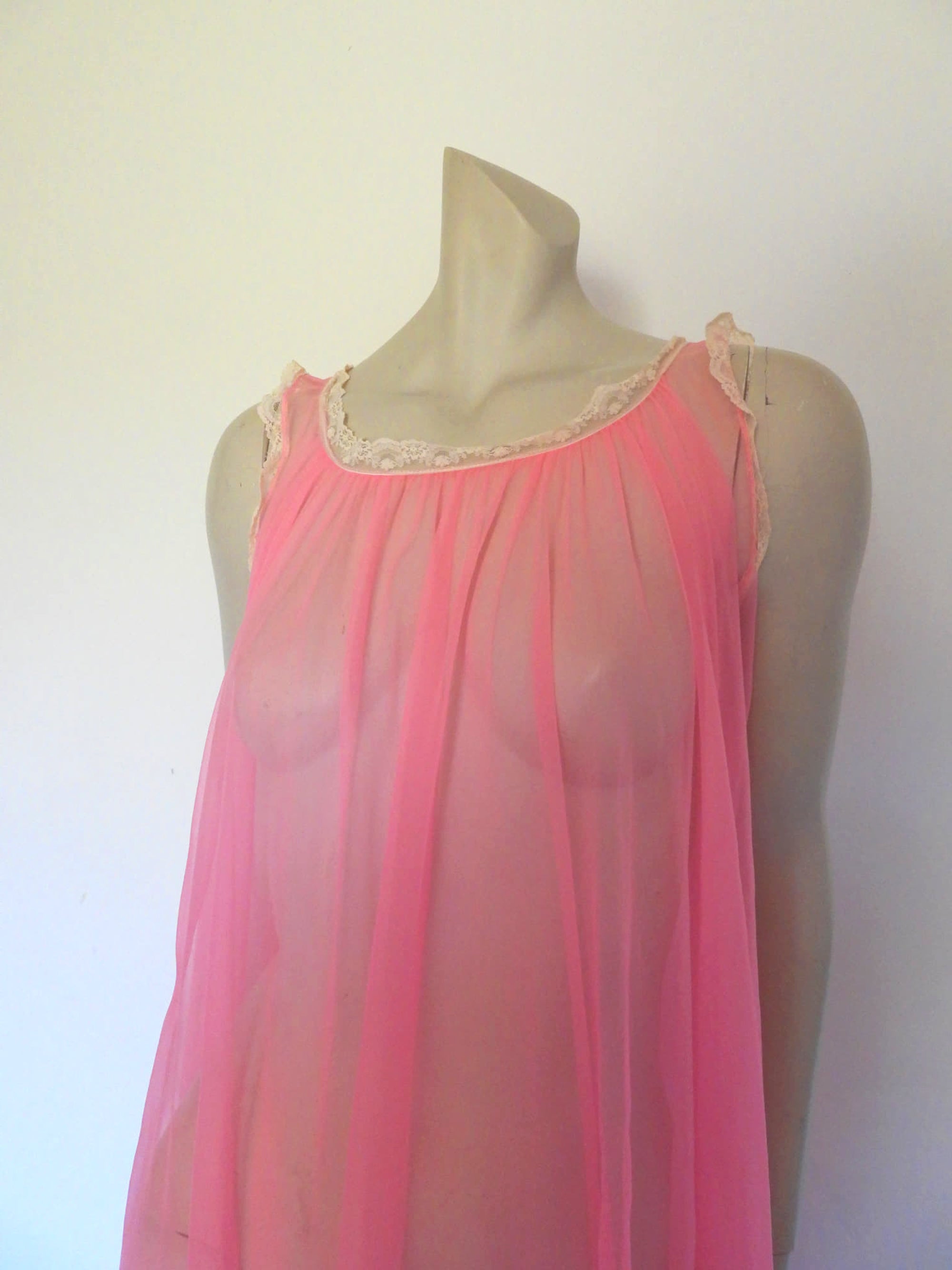 Super sexy sheer hot pink negligee nightgown. Has some issues but presents well. Very full negligee has wide neckline trimmed with cream lace. The hem is trimmed with a deep border of cream lace. There's a fixed tie at centre back which can tie in the fullness if you prefer. I have appliqued two flowers over two large holes in the front. Fabric is a single layer of sheer nylon. Made in the USA for Berkliff - 1960s. Small to Medium.