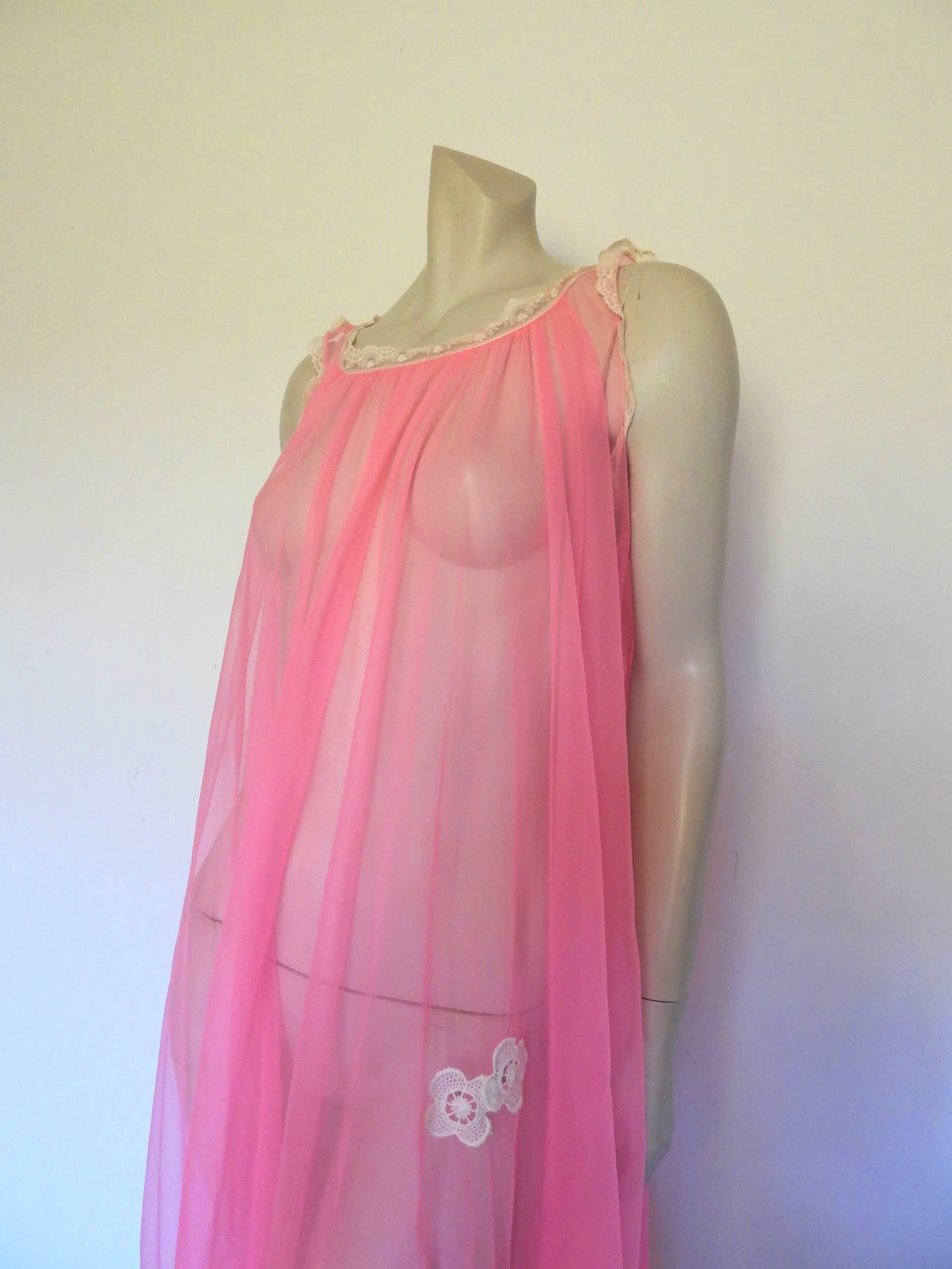 Super sexy sheer hot pink negligee nightgown. Has some issues but presents well. Very full negligee has wide neckline trimmed with cream lace. The hem is trimmed with a deep border of cream lace. There's a fixed tie at centre back which can tie in the fullness if you prefer. I have appliqued two flowers over two large holes in the front. Fabric is a single layer of sheer nylon. Made in the USA for Berkliff - 1960s. Small to Medium.