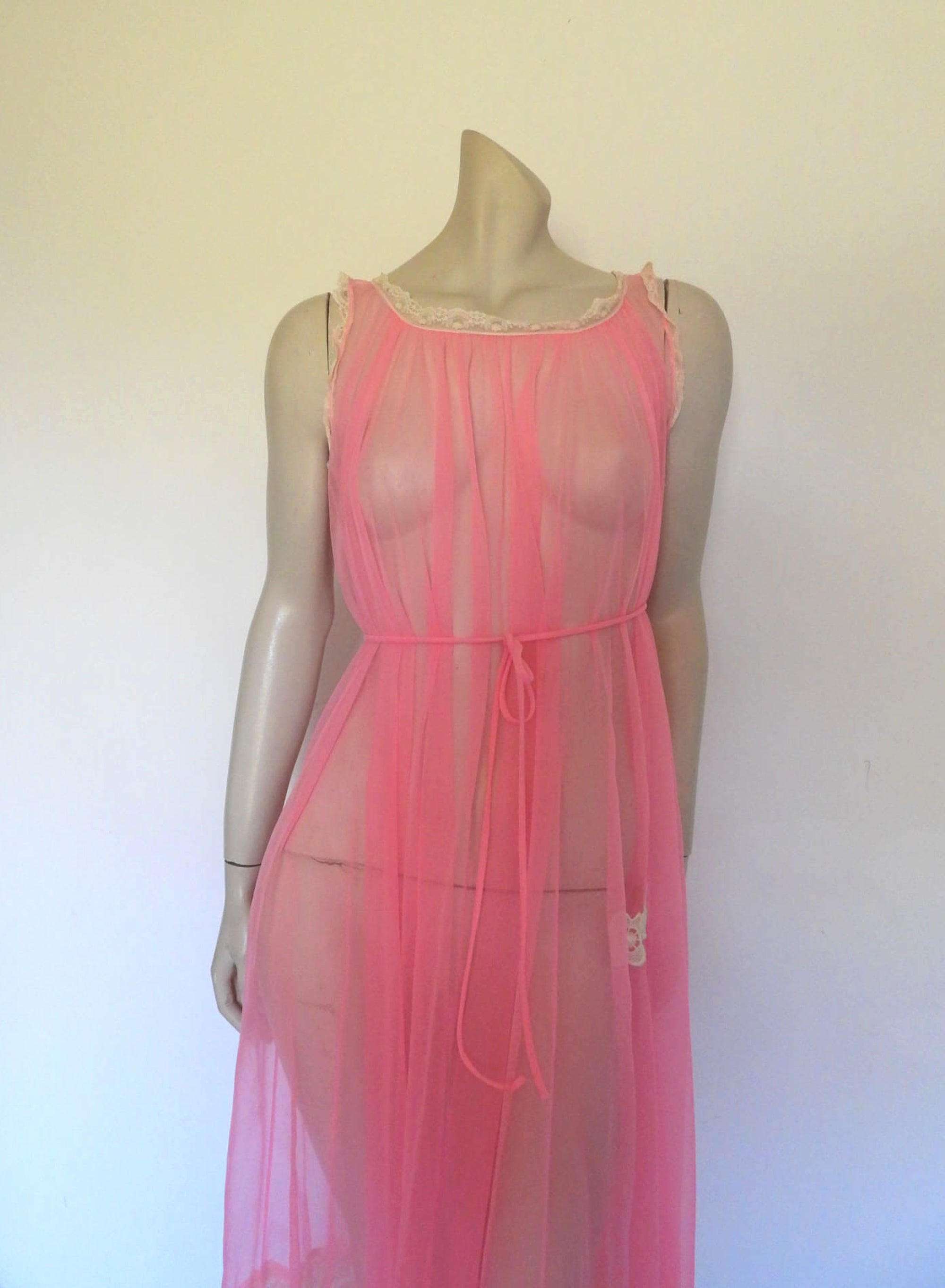 Super sexy sheer hot pink negligee nightgown. Has some issues but presents well. Very full negligee has wide neckline trimmed with cream lace. The hem is trimmed with a deep border of cream lace. There's a fixed tie at centre back which can tie in the fullness if you prefer. I have appliqued two flowers over two large holes in the front. Fabric is a single layer of sheer nylon. Made in the USA for Berkliff - 1960s. Small to Medium.
